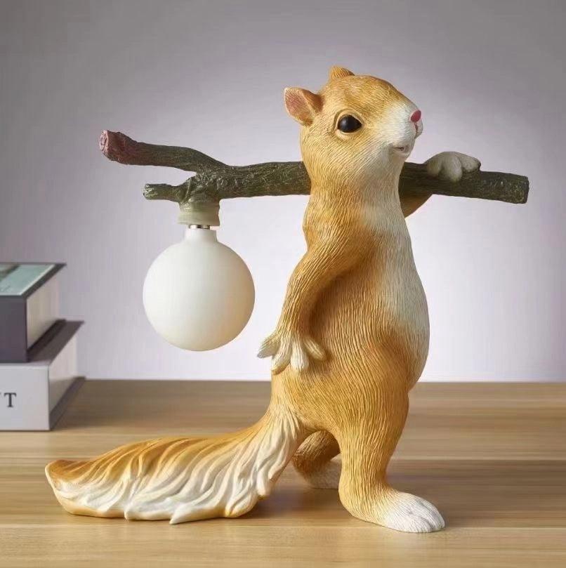 A tan-gold Clérachic squirrel lamp with an unlit frosted round bulb, on a wooden shelf with books against a lilac wall.