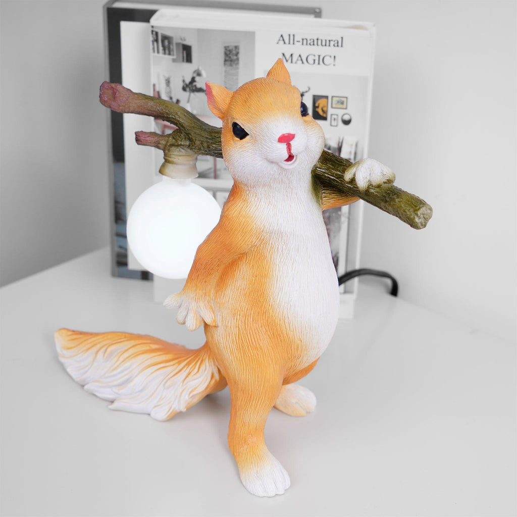 Front three-quarter view of the Clérachic squirrel lamp in orange and white, a lit round bulb hanging beside a book on white.