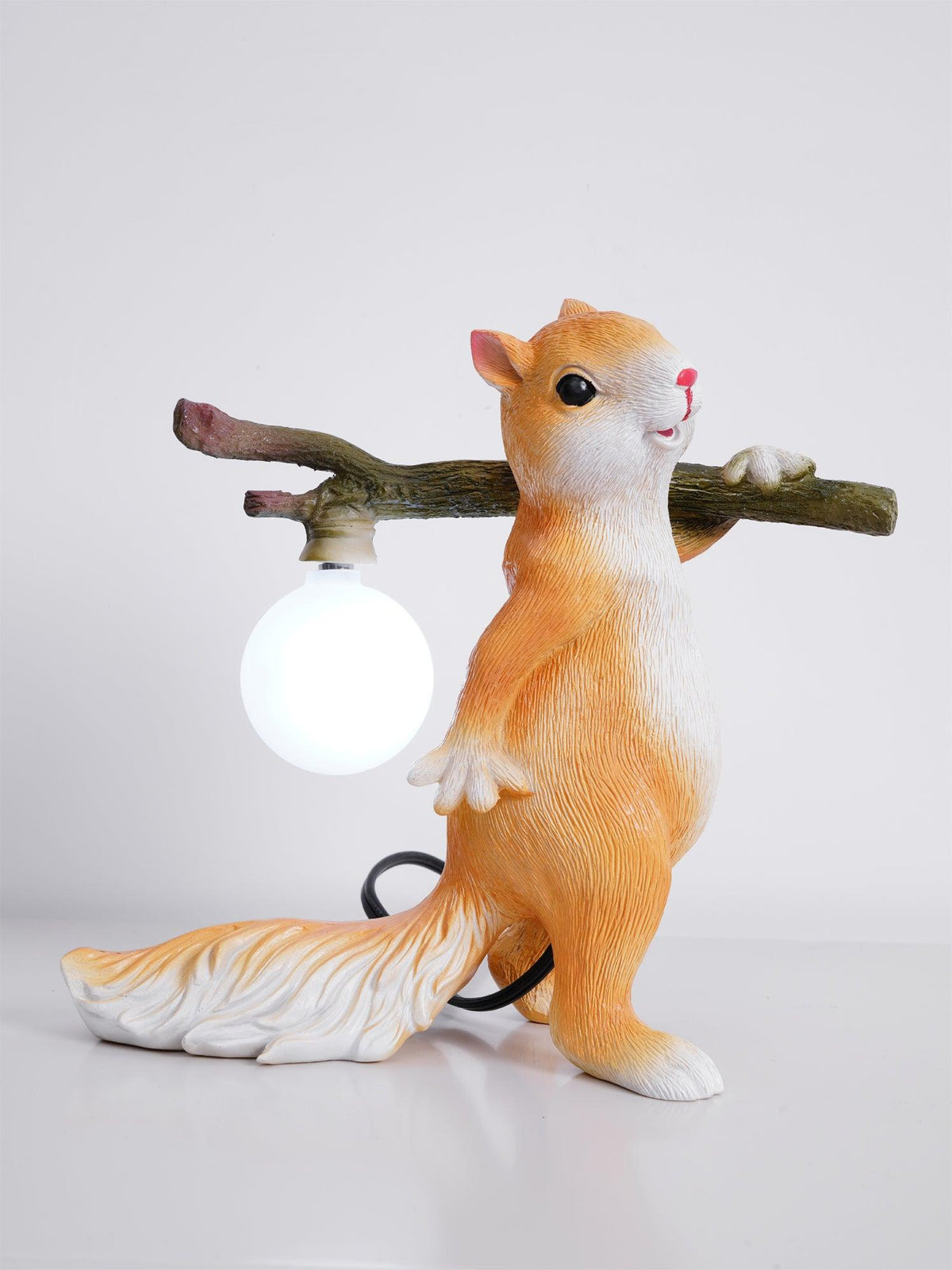 Orange and white Clérachic squirrel lamp with its round bulb lit cool white, on a pale surface with the black cable curling behind.