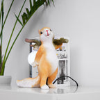 Clérachic squirrel lamp on a white shelf beside a tall green plant and a stack of books, its round bulb glowing.