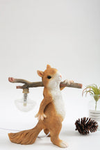 A Clérachic squirrel lamp on a white tabletop, beside a pine cone and a glass vase holding a small plant.
