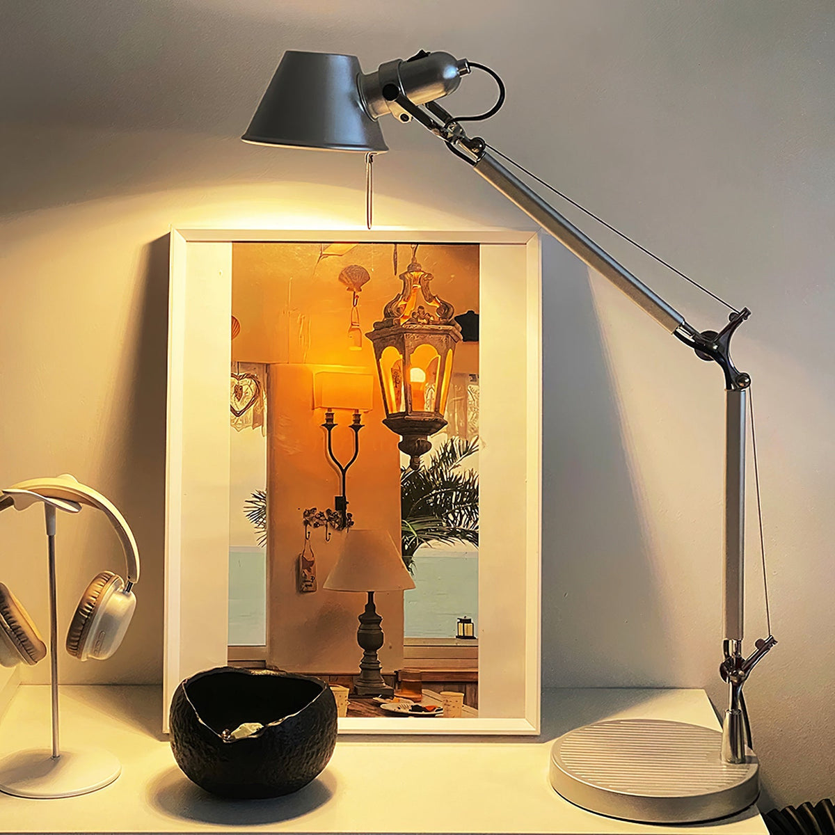 A Clérachic chrome desk lamp glowing warm against a framed picture, with a pair of headphones on a stand beside it.