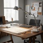 A Clérachic desk lamp reaches over a wooden drafting table covered in plans, with a pinboard behind it.