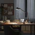A Clérachic desk lamp lit above a wooden desk with a laptop and papers, beside a pinboard and sheer curtains.