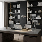 This Clérachic desk lamp stands on a grey desk with a laptop, in front of dark shelves holding vases and books.
