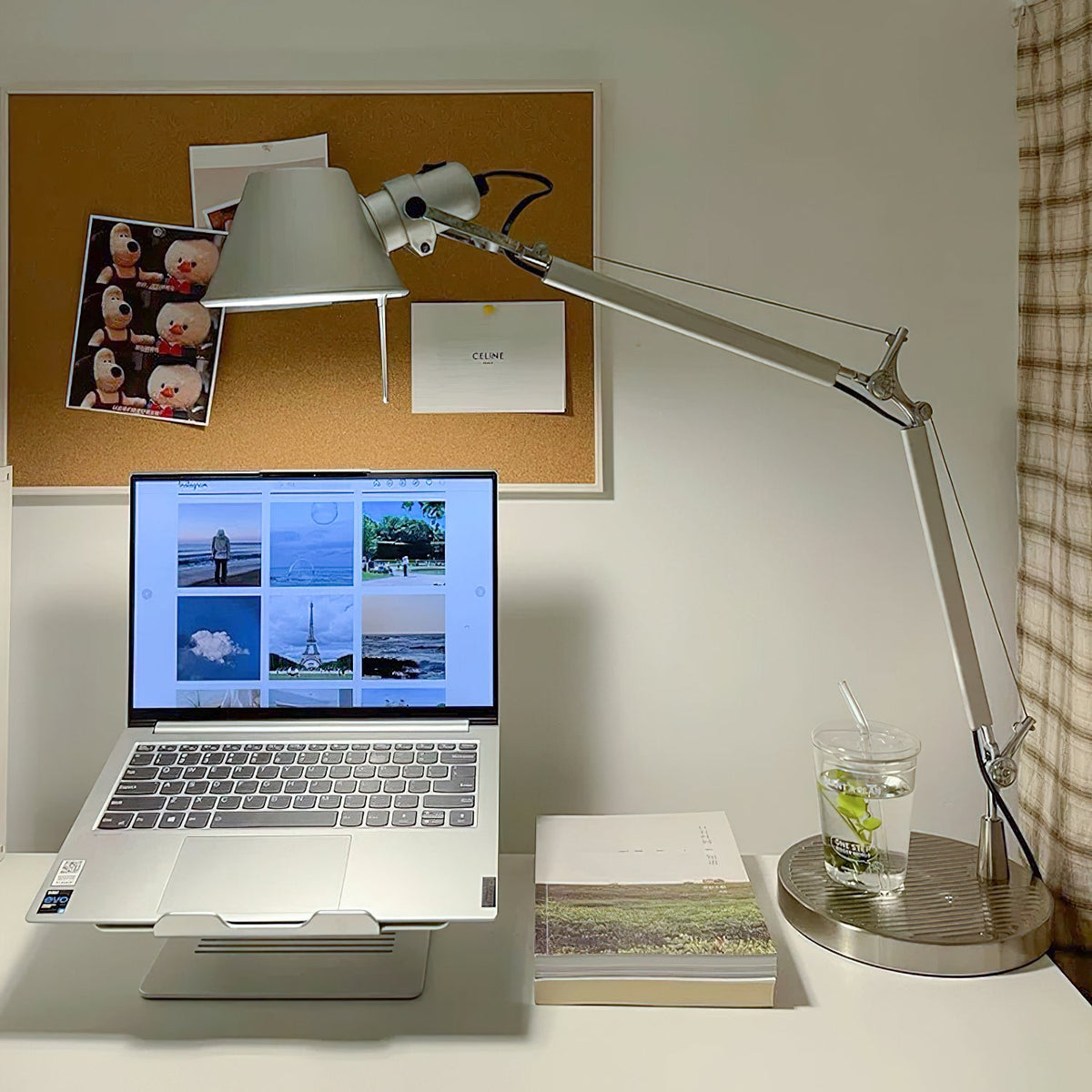 Clérachic desk lamp with its shade tilted down over a laptop on a stand, with a pinboard of photos behind.