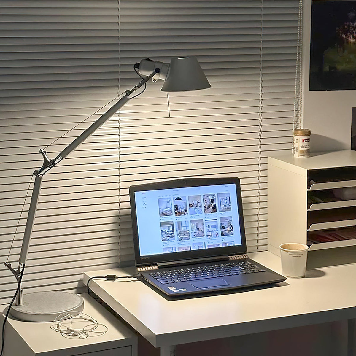 A Clérachic desk lamp lit beside a window with slatted blinds, with its arm stretching over a laptop.
