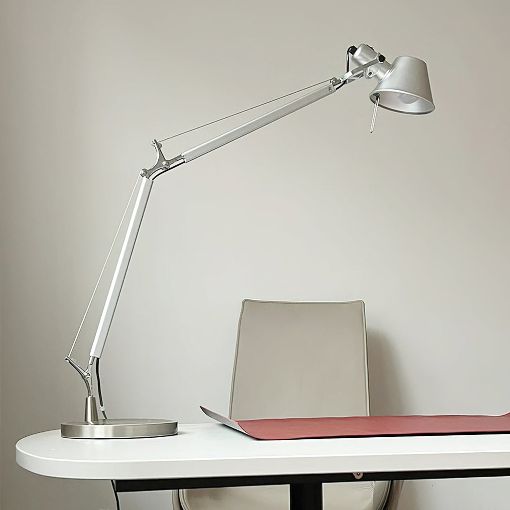 This Clérachic chrome desk lamp stands on the edge of a white desk, with a red desk mat and a pale chair behind.