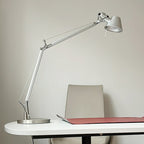 This Clérachic chrome desk lamp stands on the edge of a white desk, with a red desk mat and a pale chair behind.