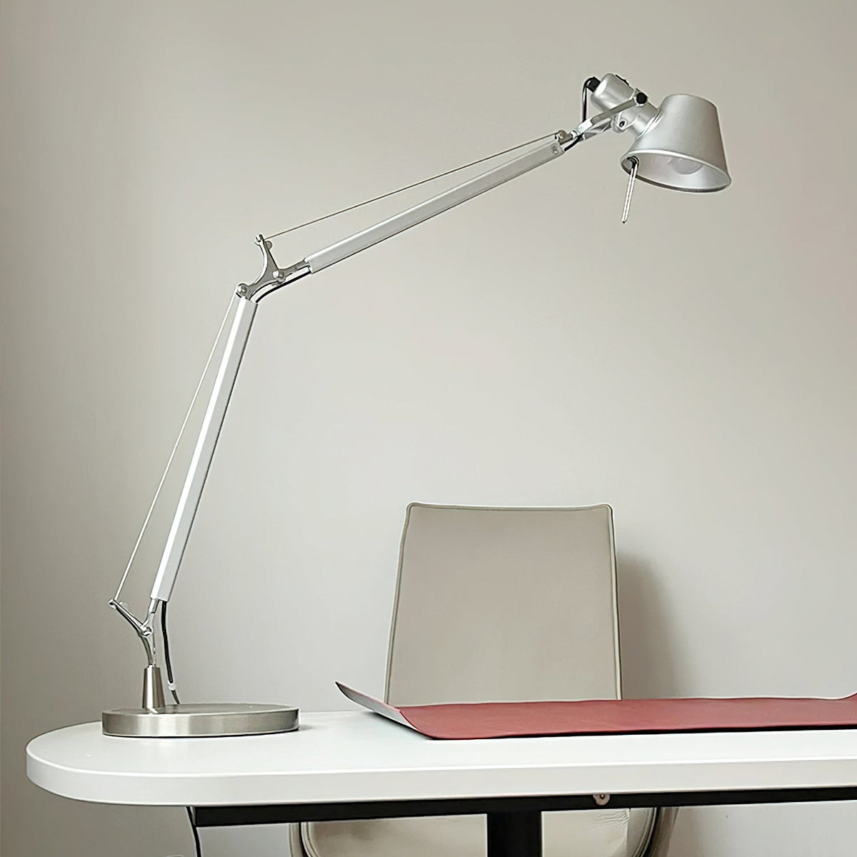 This Clérachic chrome desk lamp stands on the edge of a white desk, with a red desk mat and a pale chair behind.