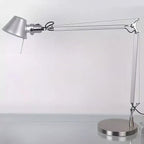 A Clérachic desk lamp with its arm extended, standing on a pale desk against a soft grey wall.