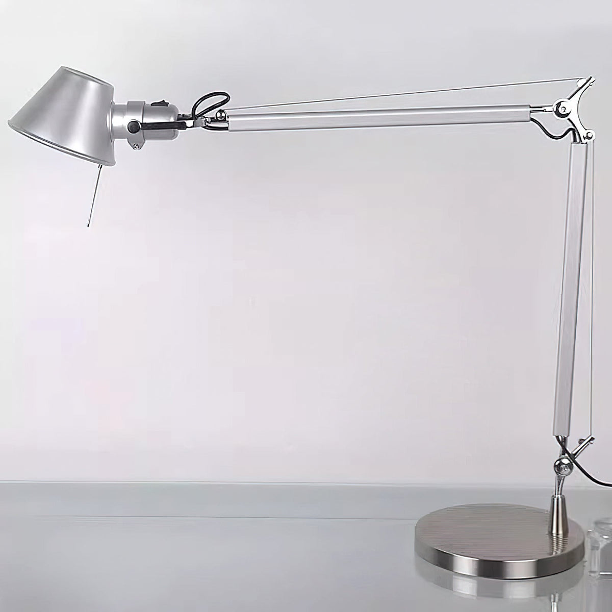 A Clérachic desk lamp with its arm extended, standing on a pale desk against a soft grey wall.