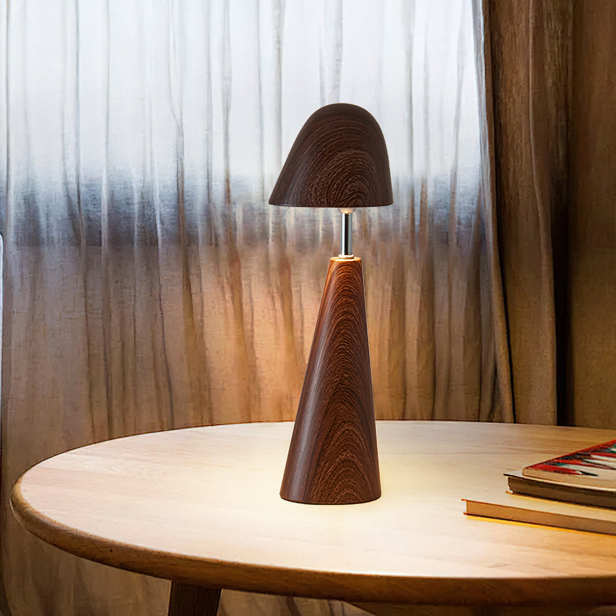 Lit Clérachic table lamp in walnut on a round wooden table, its dome shade glowing warm in front of sheer curtains.