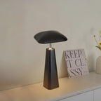 Black Clérachic table lamp with a flat, rounded-square shade and a tapered base, glowing on a white sideboard beside a printed card.