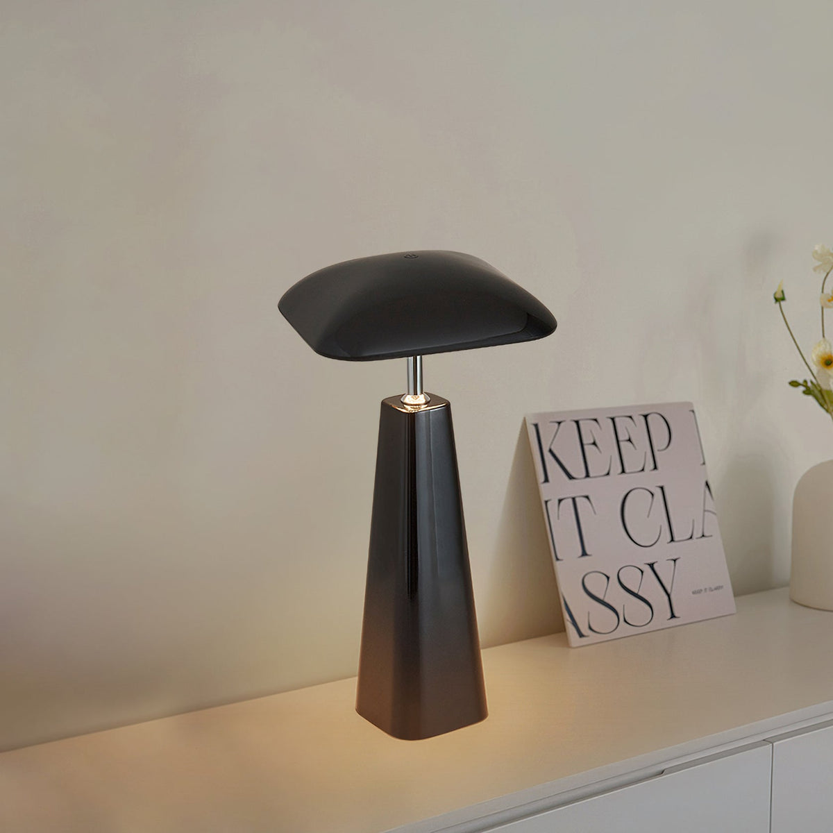Black Clérachic table lamp with a flat, rounded-square shade and a tapered base, glowing on a white sideboard beside a printed card.