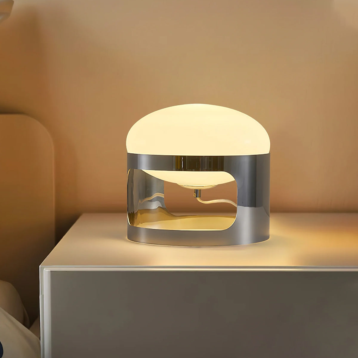 This Clérachic table lamp glows warm on a white bedside unit, its opal dome sitting over a chrome band with a curved cutout.