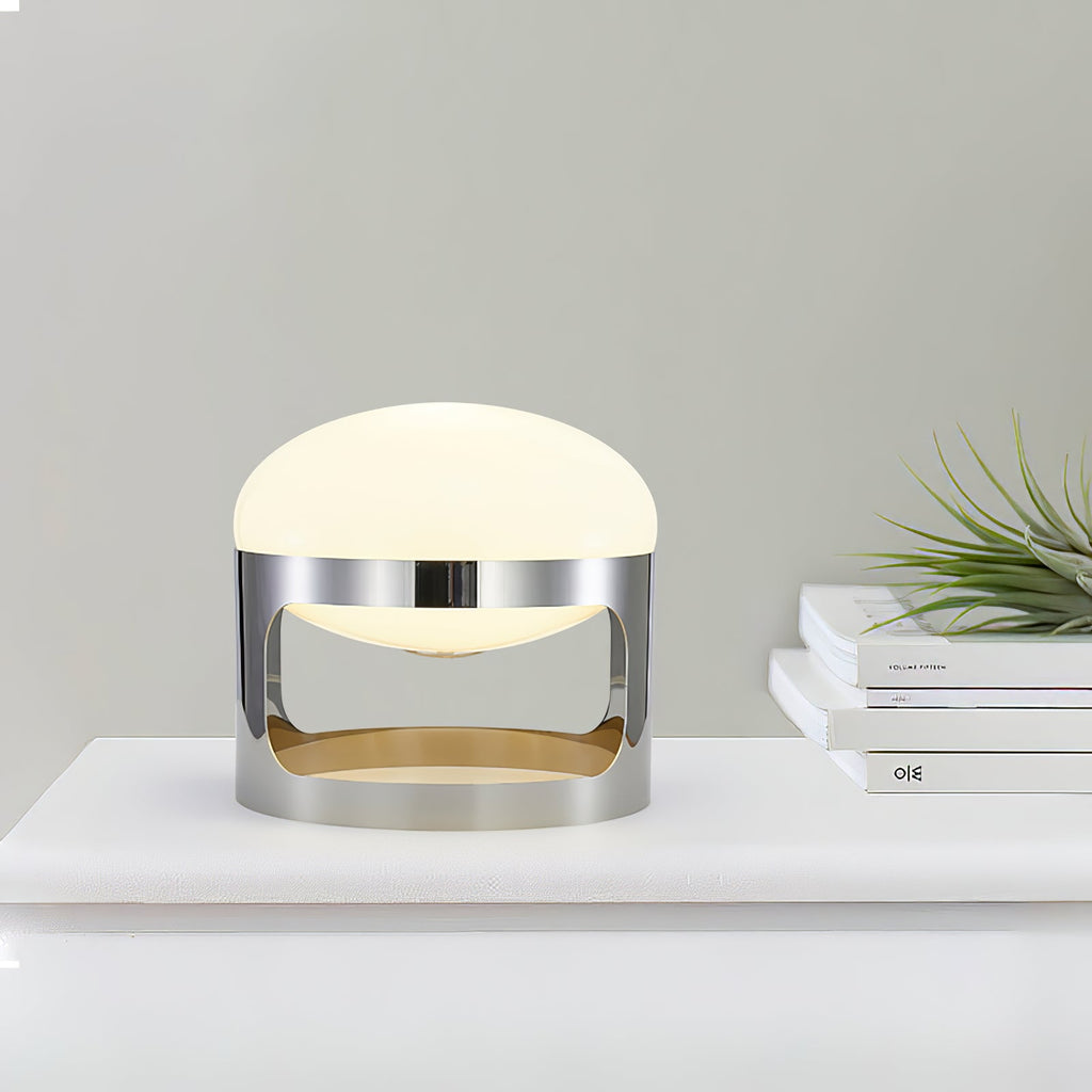 A lit Clérachic table lamp on a white shelf beside a stack of books and a leafy plant, against a pale green-grey wall.