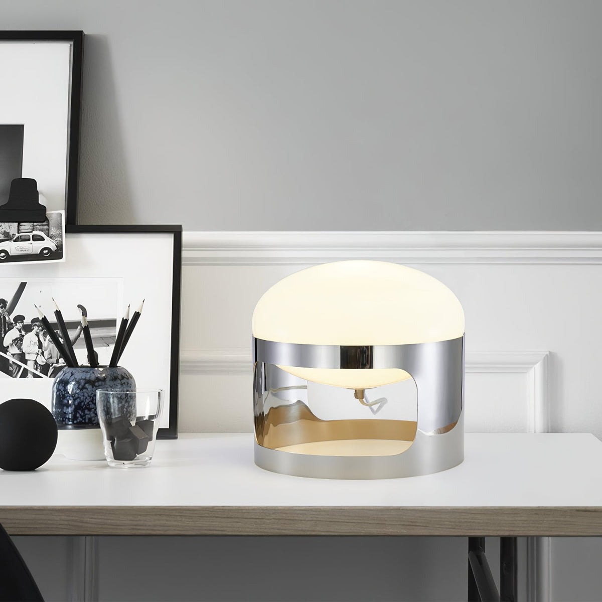 Clérachic table lamp with a chrome cutout band on a white desk, next to a framed photo, a pot of pencils and a glass.