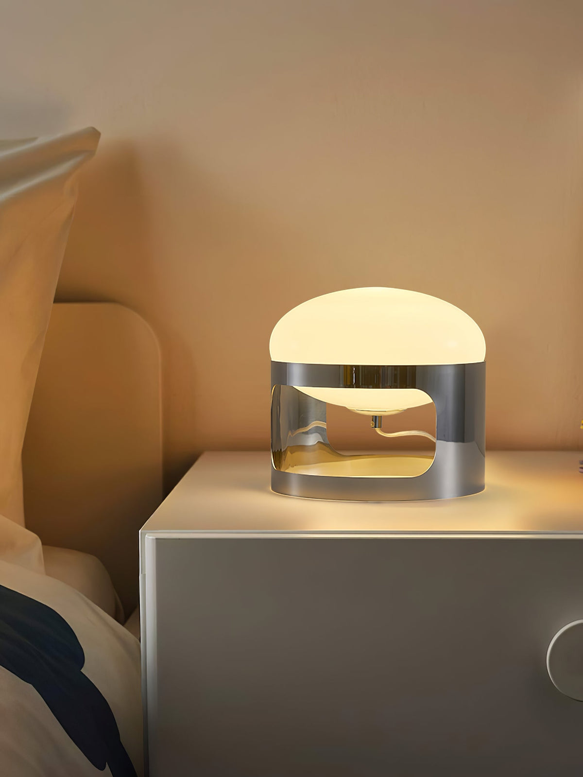 A Clérachic table lamp glows on a pale bedside unit in a warm-lit bedroom, with a pillow just visible at the edge.