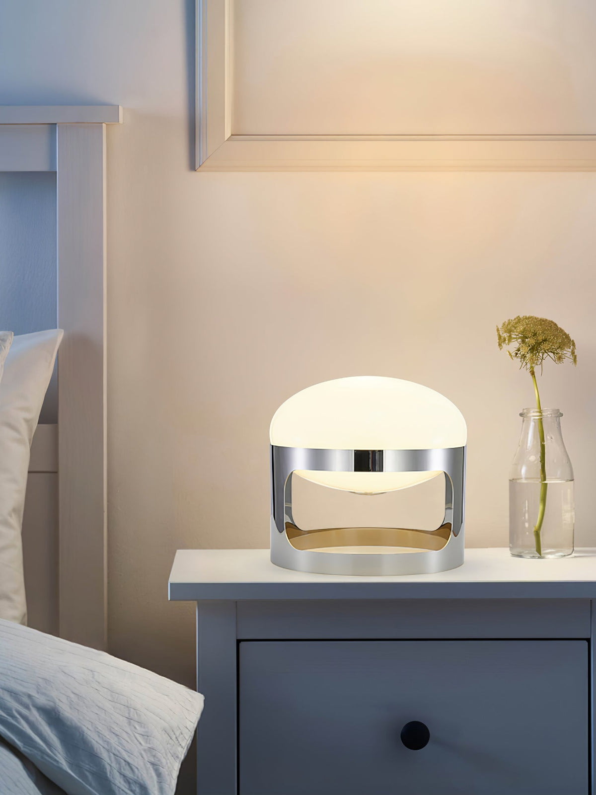 A Clérachic table lamp lit on a navy bedside chest, with a stem in a glass bottle and a soft glow on the wall behind.