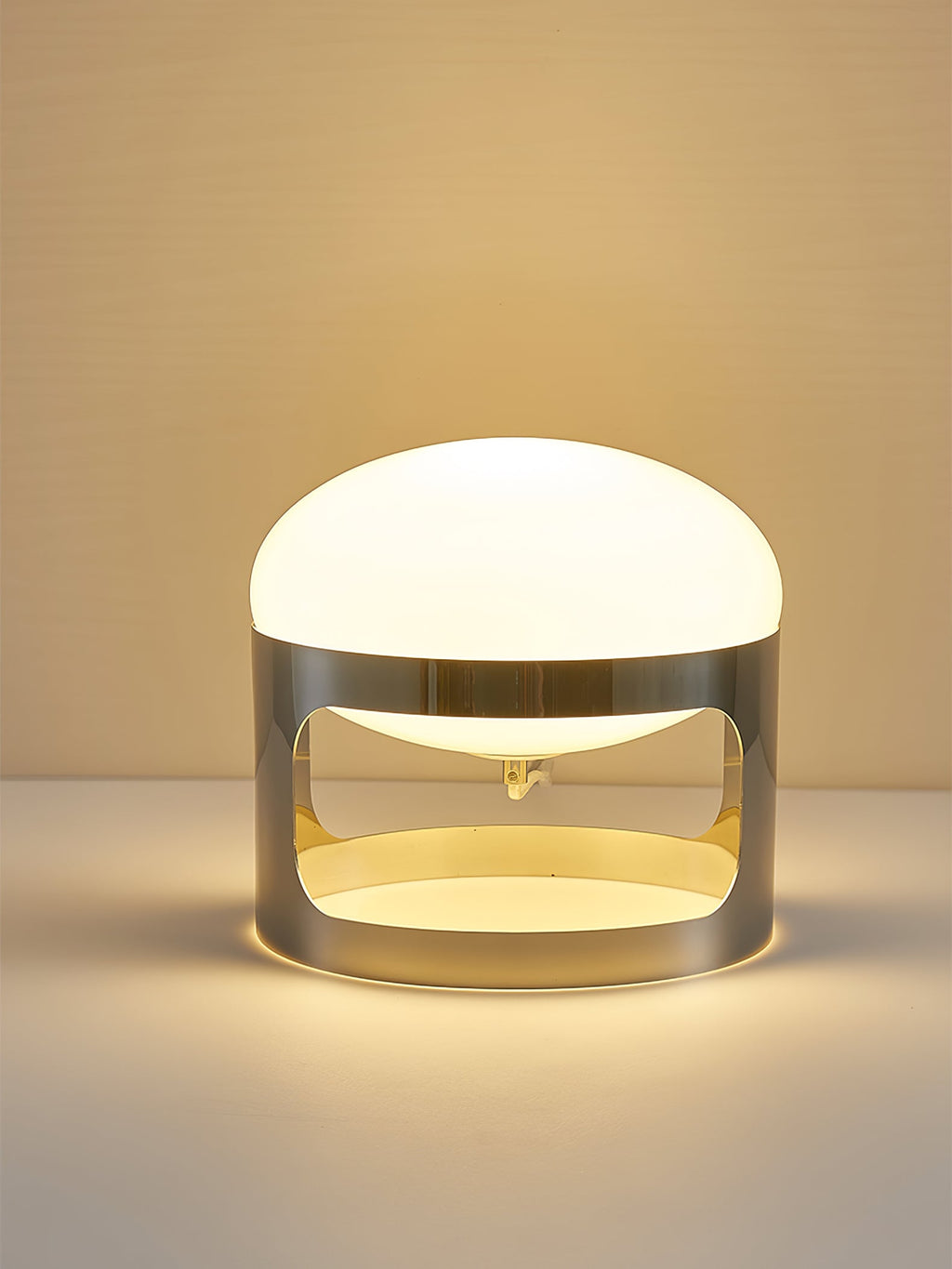 A Clérachic table lamp lit in warm light on a pale amber backdrop, with a gold-toned glow inside the chrome band.