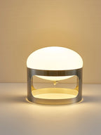 A Clérachic table lamp lit in warm light on a pale amber backdrop, with a gold-toned glow inside the chrome band.