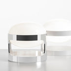 A chrome Clérachic table lamp in the foreground, with a white-finish version of the same shape softly blurred behind it.