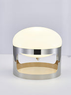 Clérachic table lamp lit on a pale grey backdrop, with its opal dome above a chrome band that is open at the sides.
