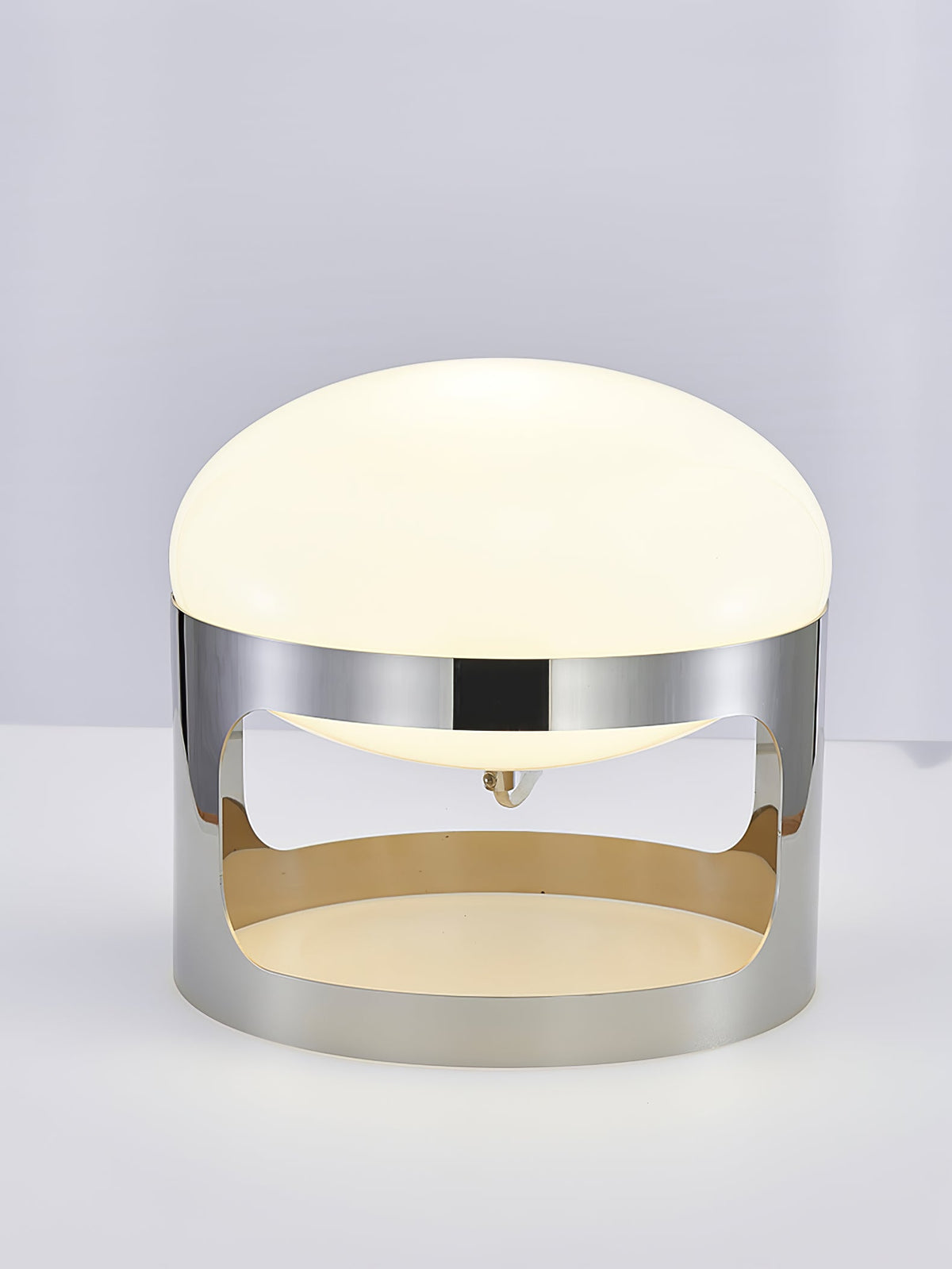 Clérachic table lamp lit on a pale grey backdrop, with its opal dome above a chrome band that is open at the sides.