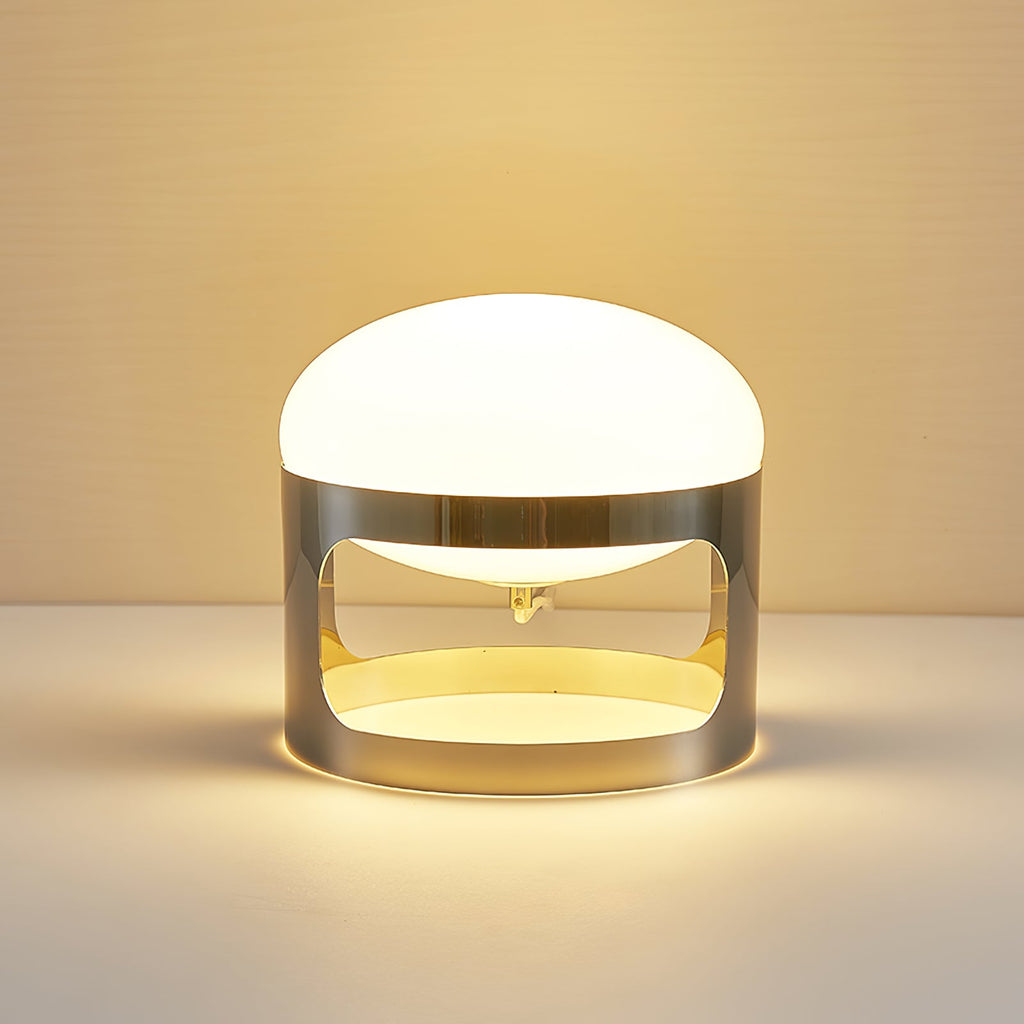 A Clérachic table lamp lit on a light surface in warm light, with a gold glow inside the chrome band.