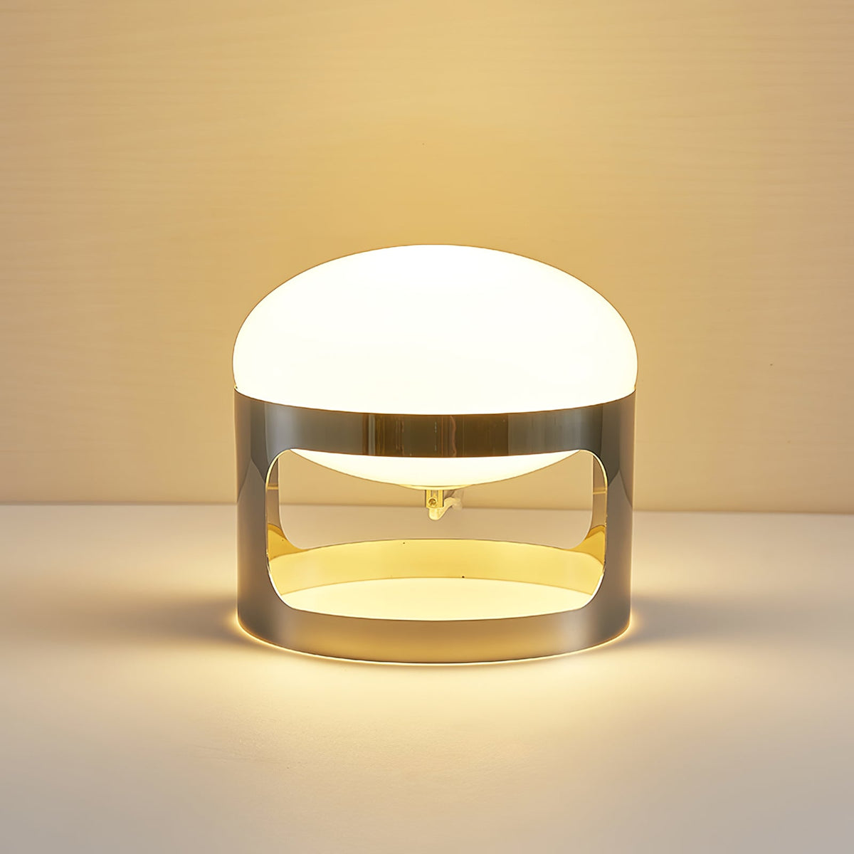 A Clérachic table lamp lit on a light surface in warm light, with a gold glow inside the chrome band.
