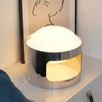 Close angled view of a lit Clérachic table lamp on a white chest, the opal dome glowing and the chrome band cutout showing warm light.