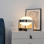 A lit Clérachic table lamp on a white drawer chest beside a blue sofa arm, with a line-drawn print behind it.