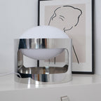 A Clérachic table lamp, switched off, on a white drawer chest, with its polished chrome band reflecting the room and a framed line drawing behind it.