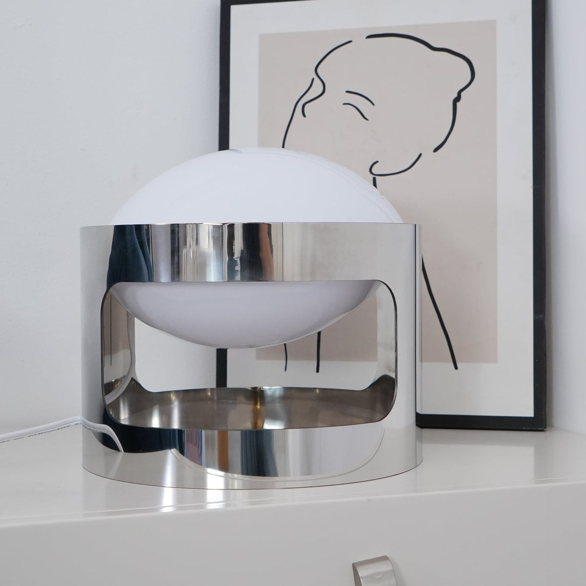 A Clérachic table lamp, switched off, on a white drawer chest, with its polished chrome band reflecting the room and a framed line drawing behind it.