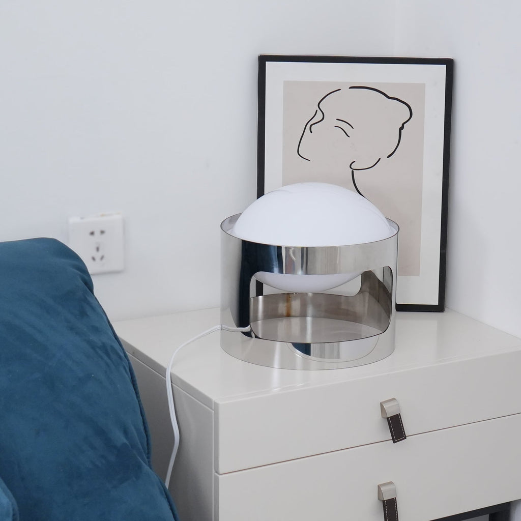 A Clérachic table lamp unlit on a white chest beside a blue sofa, with a wall socket and framed line drawing behind.