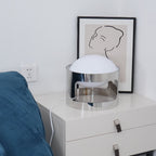 A Clérachic table lamp unlit on a white chest beside a blue sofa, with a wall socket and framed line drawing behind.