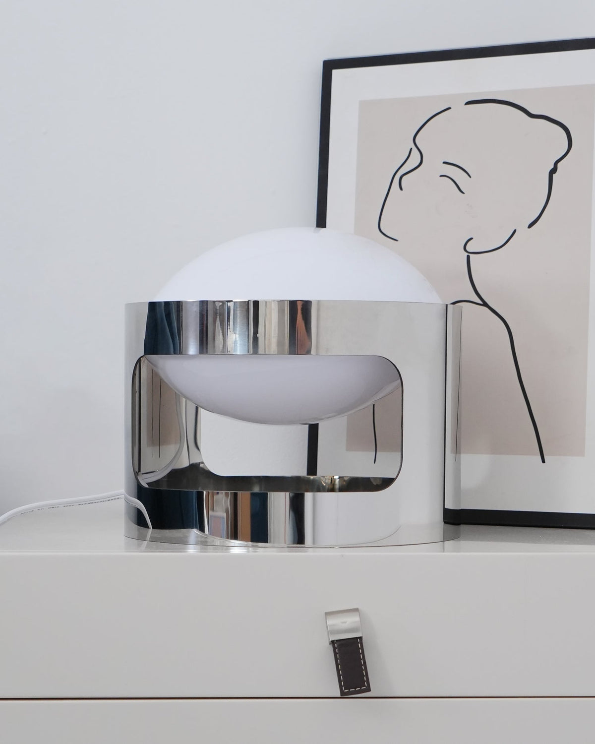 Clérachic table lamp, switched off, in front of a framed line drawing, its chrome band cutout showing the opal bowl inside.