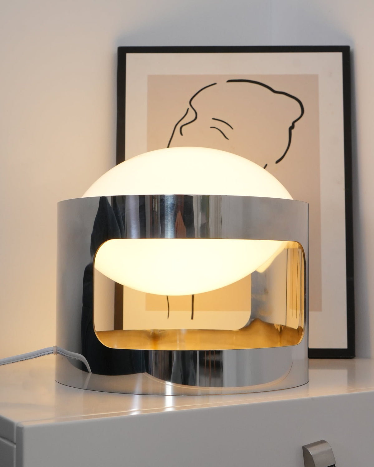 A Clérachic table lamp lit on a white chest, the chrome band cutout showing a warm glow beneath the white dome.