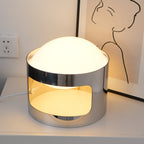 Clérachic table lamp lit on a white bedside chest next to a wall socket, with a line-drawn print behind and the cord trailing left.