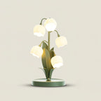 Clérachic table lamp with five glowing bell shades on green stems, with leaf-shaped metal blades, on a soft cream backdrop.