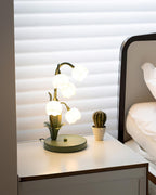 A Clérachic table lamp lit on a white bedside table with a small cactus, beside a bed and window blinds.