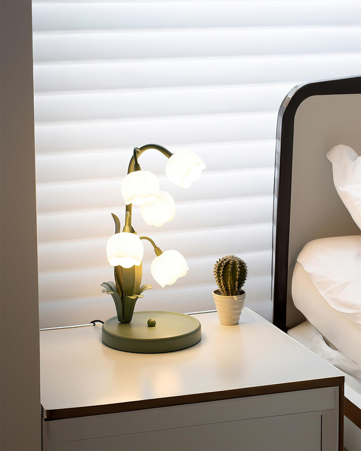 A Clérachic table lamp lit on a white bedside table with a small cactus, beside a bed and window blinds.