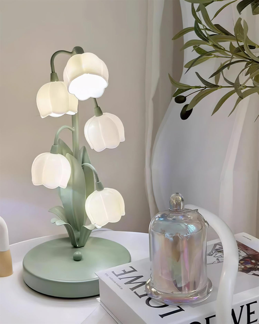This Clérachic table lamp sits on a white side table beside a glass cloche and a magazine, with olive branches and sheer curtains behind.