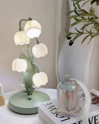 This Clérachic table lamp sits on a white side table beside a glass cloche and a magazine, with olive branches and sheer curtains behind.