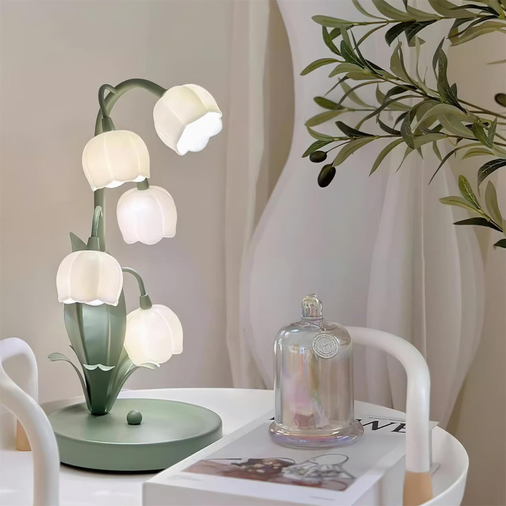 A Clérachic table lamp lit next to a glass cloche on a magazine, with olive leaves and a pale curtain in the background.