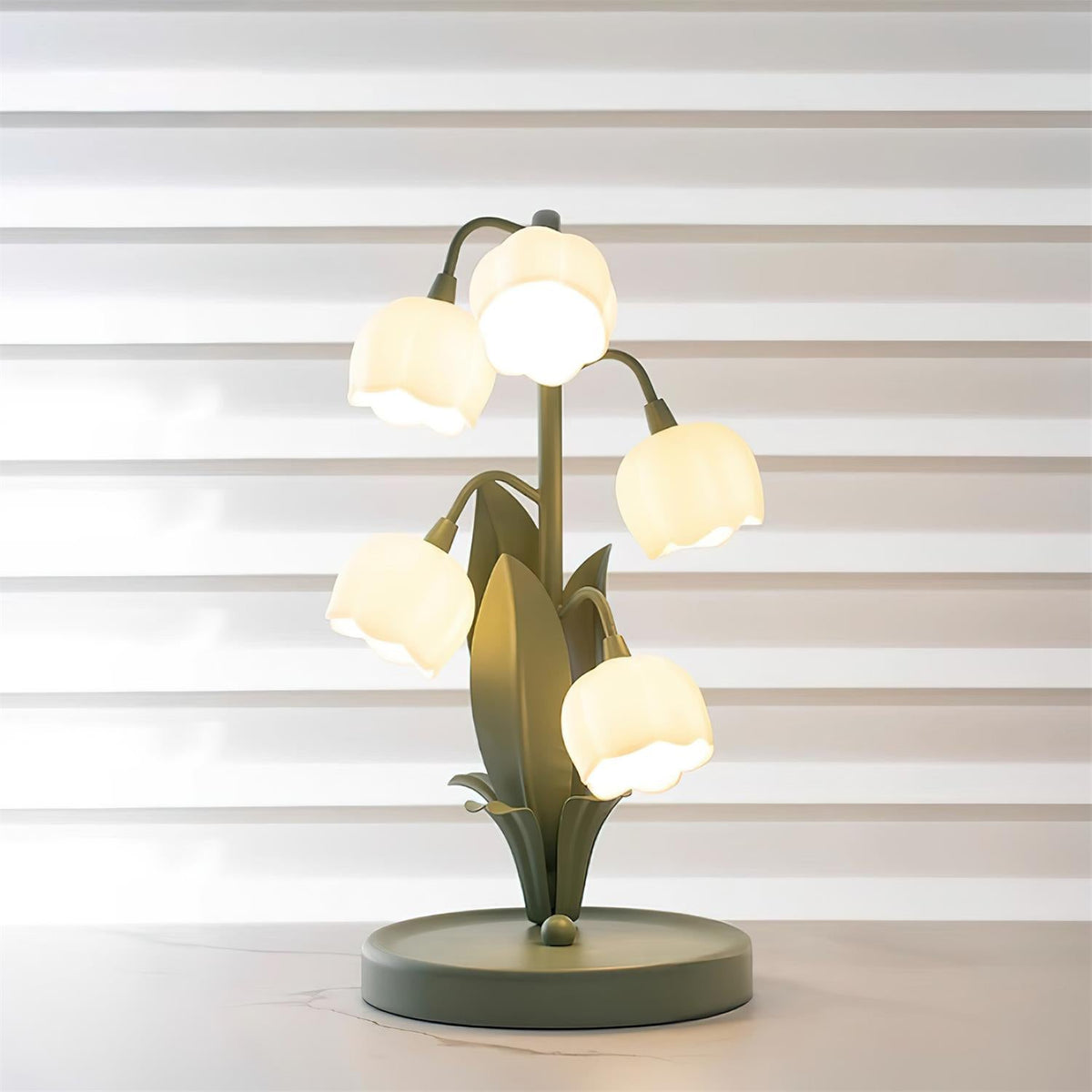 A lit Clérachic table lamp on a white marble surface, with its bell shades glowing against striped window blinds.