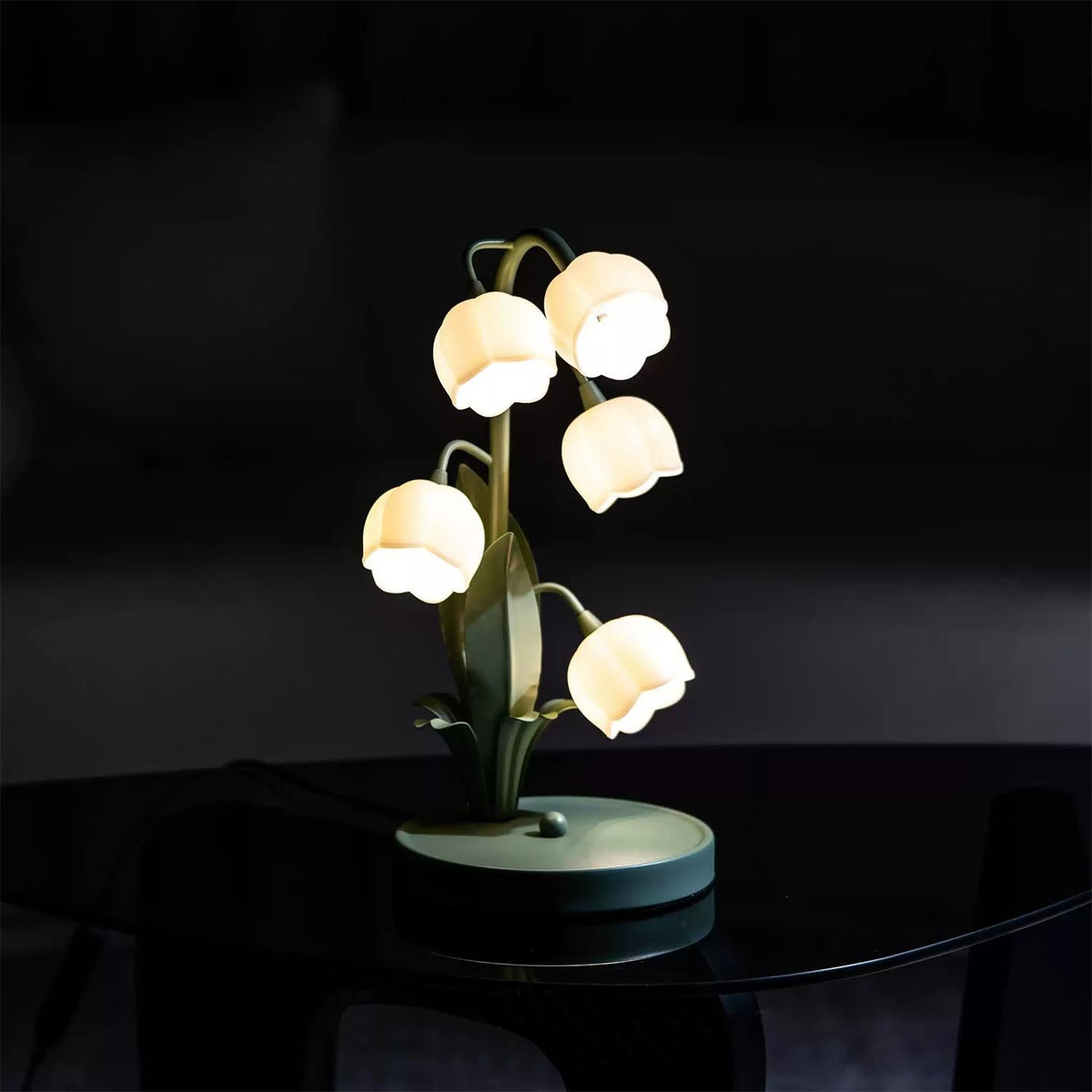 This Clérachic table lamp glows in a dark room, standing on a dark glass tabletop with its green base just visible.