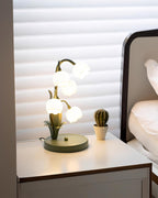 A Clérachic table lamp lit on a white bedside chest with a small cactus, a bed and window blinds in the background.