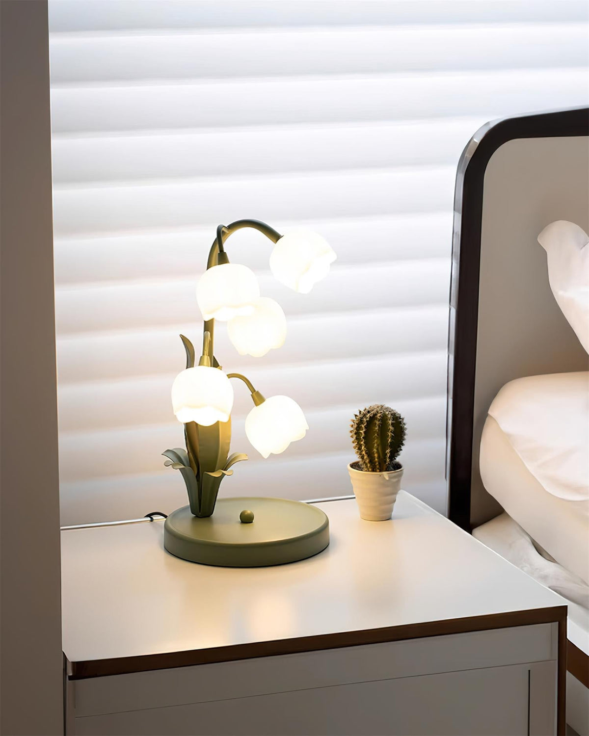 A Clérachic table lamp lit on a white bedside chest with a small cactus, a bed and window blinds in the background.